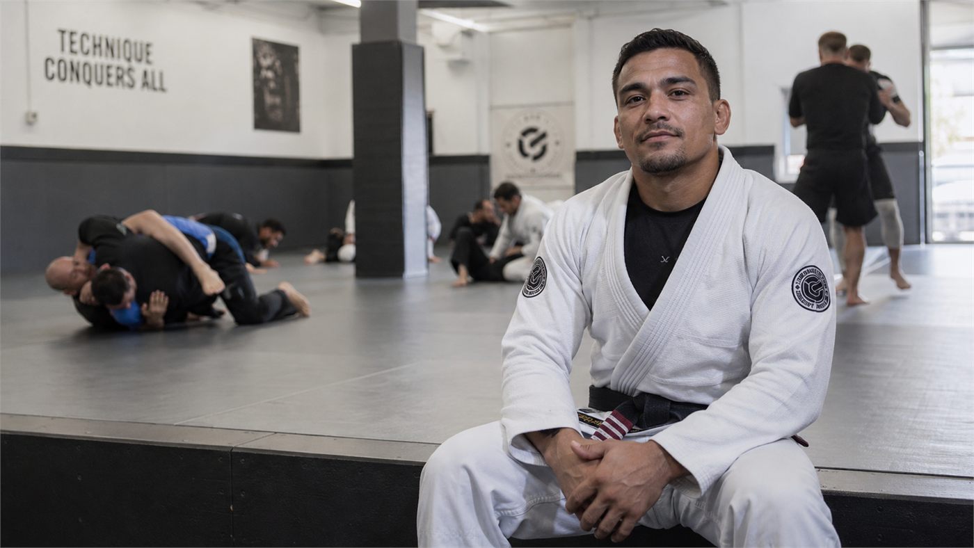An instructor sitting on the mat while a class trains behind him.